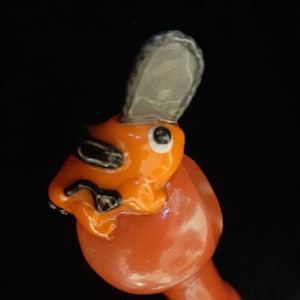 Puede incluir: Figurilla de cerámica naranja y negra de una rana con un sombrero gris. La rana está sentada sobre una base roja.