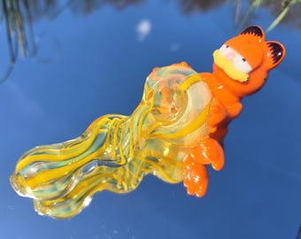 May include: A glass pipe with a yellow and green swirl design. The pipe is shaped like a wave and has a cartoon orange cat character on the end.