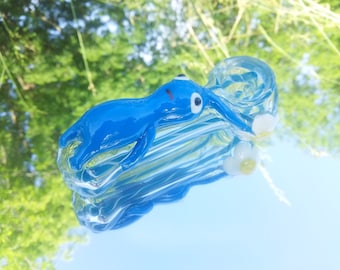 Può includere: Un  pipa di vetro blu a forma di personaggio dei cartoni animati con un motivo a fiore bianco. La pipa è realizzata in vetro trasparente con accenti blu.