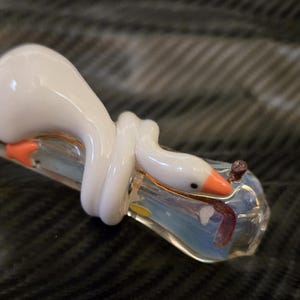 May include: A glass pipe with a white goose design. The goose has an orange beak and is wrapped around the clear glass pipe. The pipe has a colorful, iridescent appearance.