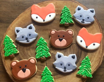 Puede incluir: Una colección de nueve galletas de azúcar decoradas en una tabla de madera. Las galletas tienen forma de osos, zorros, mapaches y pinos. Las galletas están decoradas con glaseado verde, naranja, gris y marrón.