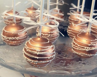 Puede incluir: Primer plano de un plato de cake pops decorados con glaseado blanco y chispas blancas. Los cake pops están en palitos blancos y tienen una base de color cobre.