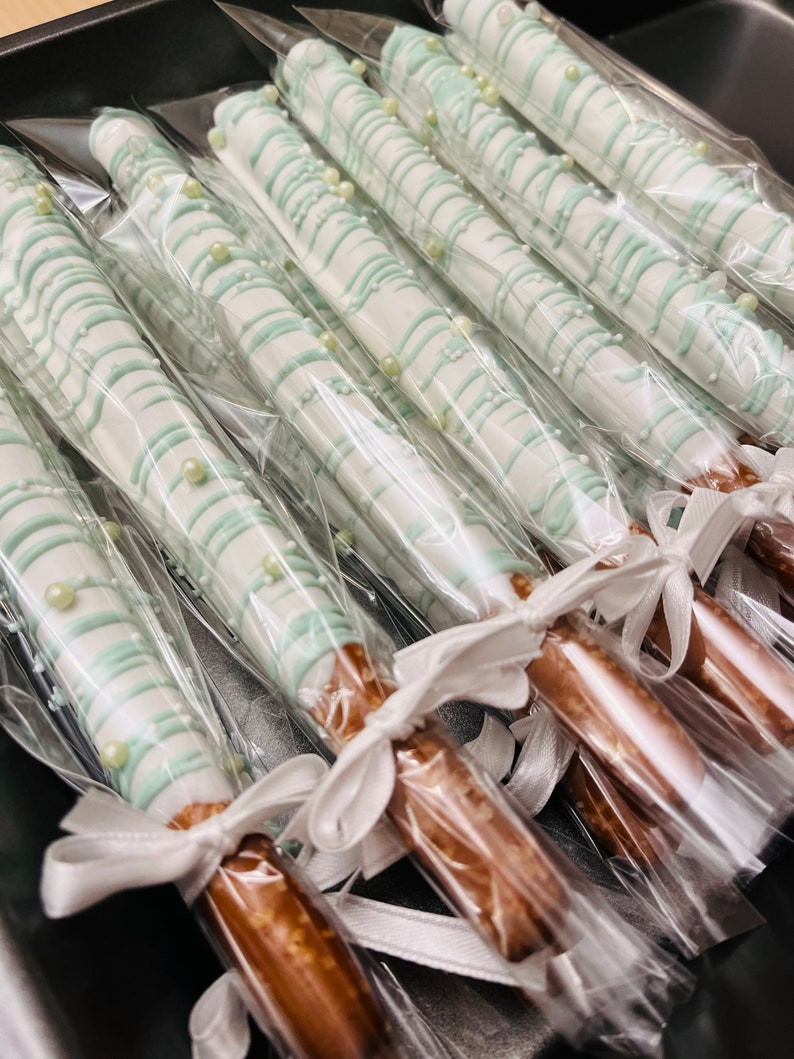 Puede incluir: Primer plano de varios bastones de pretzel cubiertos de chocolate blanco y decorados con chispas verdes y blancas. Los pretzels est&aacute;n envueltos individualmente en bolsas de pl&aacute;stico transparentes y atados con cintas blancas.