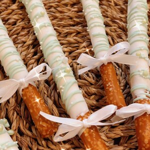 Puede incluir: Primer plano de cuatro palitos de pretzel cubiertos de chocolate blanco y decorados con rayas verdes y blancas y chispas blancas. Los pretzels est&aacute;n atados con lazos de cinta blanca.