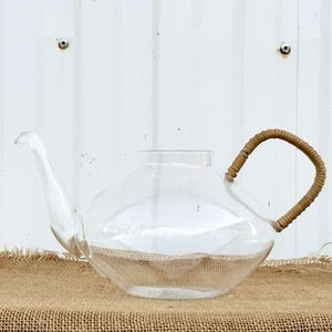 Può includere: Una teiera di vetro trasparente con un manico intrecciato marrone. La teiera è appoggiata su una superficie di juta marrone.