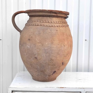 以下が含まれることがあります： 口が広く、持ち手が1つ付いた、大きな茶色のテラコッタ製の壺。壺の上部には装飾的な帯があり、少し風化している。