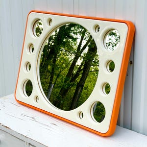 May include: Square mirror with a cream-colored frame and an orange border. The mirror has a large central circular mirror and smaller circular mirrors around the perimeter. The reflection shows a green forest scene. A unique decorative piece.