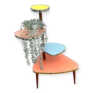 Può includere: Un portapiante a più livelli dal design retrò. Il supporto presenta tre ripiani triangolari nei colori corallo, azzurro e giallo, sostenuti da una struttura in legno. Una pianta ricadente in un vaso bianco si trova sul ripiano centrale.