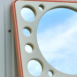 May include: A retro-style mirror with a square shape and rounded corners. The frame is a light peach color, surrounding a cream-colored interior with several circular mirror cutouts. The main mirror reflects a blue sky with clouds.