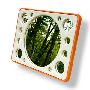 May include: Square mirror with an orange frame and a white interior. The mirror has a circular center and several smaller circular cutouts around the perimeter. The reflection shows a green forest scene.
