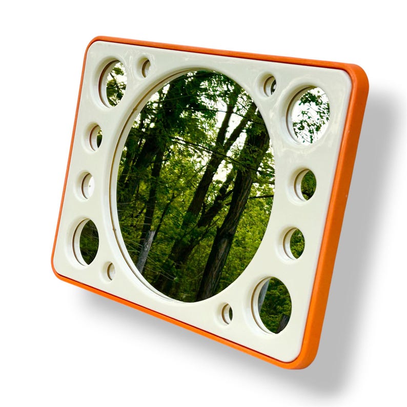 May include: Square mirror with an orange frame and a white interior. The mirror has a circular center and several smaller circular cutouts around the perimeter. The reflection shows a green forest scene.