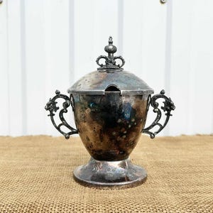 以下が含まれることがあります： 蓋と装飾的なハンドルが付いた、アンティークの銀メッキの砂糖入れ。ボウルは台座ベースと蓋の装飾的なつまみがあります。ハンドルはスクロールのようなパターンで複雑に設計されています。砂糖入れはジュートの表面に置かれています。