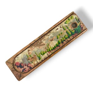 Op de afbeelding: Een vintage houten potlooddoos met een geschilderde landschapsafbeelding. De doos is rechthoekig met een schuifdeksel. Het landschap toont bomen, gebouwen en een verscheidenheid aan kleuren, waaronder groen, roze en goud.
