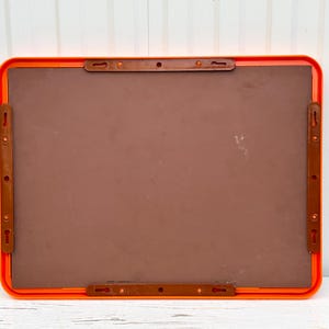 May include: A rectangular, orange-framed board with a brown interior. The back of the board features four brown plastic mounting brackets, each with multiple holes for hanging. The board is designed for wall mounting.