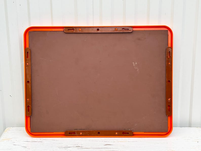 May include: A rectangular, orange-framed board with a brown interior. The back of the board features four brown plastic mounting brackets, each with multiple holes for hanging. The board is designed for wall mounting.