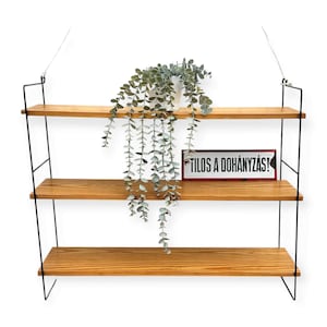 May include: A three-tiered wooden shelf with black metal supports, suspended from the ceiling. A white pot with trailing greenery sits on the top shelf. A sign that reads "TILOS A DOHÁNYZÁS!" is on the middle shelf.