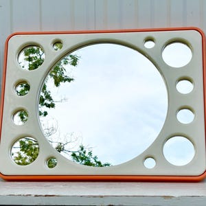 May include: Rectangular mirror with a cream-colored frame and an orange border. The mirror has a large central circular mirror and smaller circular mirrors around the perimeter. The reflection shows a sky and tree branches.