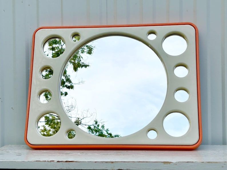 May include: Rectangular mirror with a cream-colored frame and an orange border. The mirror has a large central circular mirror and smaller circular mirrors around the perimeter. The reflection shows a sky and tree branches.