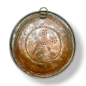 May include: A copper colander with a silver rim and a hanging loop. The colander features drainage holes in the base. The copper has an aged patina. This antique kitchenware item is likely from a bygone era.