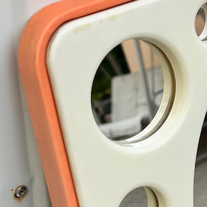 May include: Close-up of a retro-style, off-white and orange mirror. The mirror has a rounded rectangular shape with circular cutouts and an orange border. The mirror reflects a blurred outdoor scene.