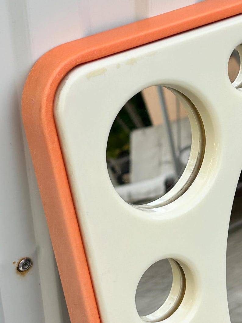 May include: Close-up of a retro-style, off-white and orange mirror. The mirror has a rounded rectangular shape with circular cutouts and an orange border. The mirror reflects a blurred outdoor scene.