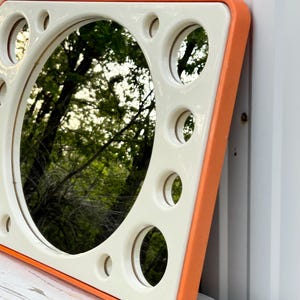 May include: A square mirror with a cream-colored frame and an orange border. The frame has several circular cutouts around the central mirror. The mirror reflects a view of trees and foliage.