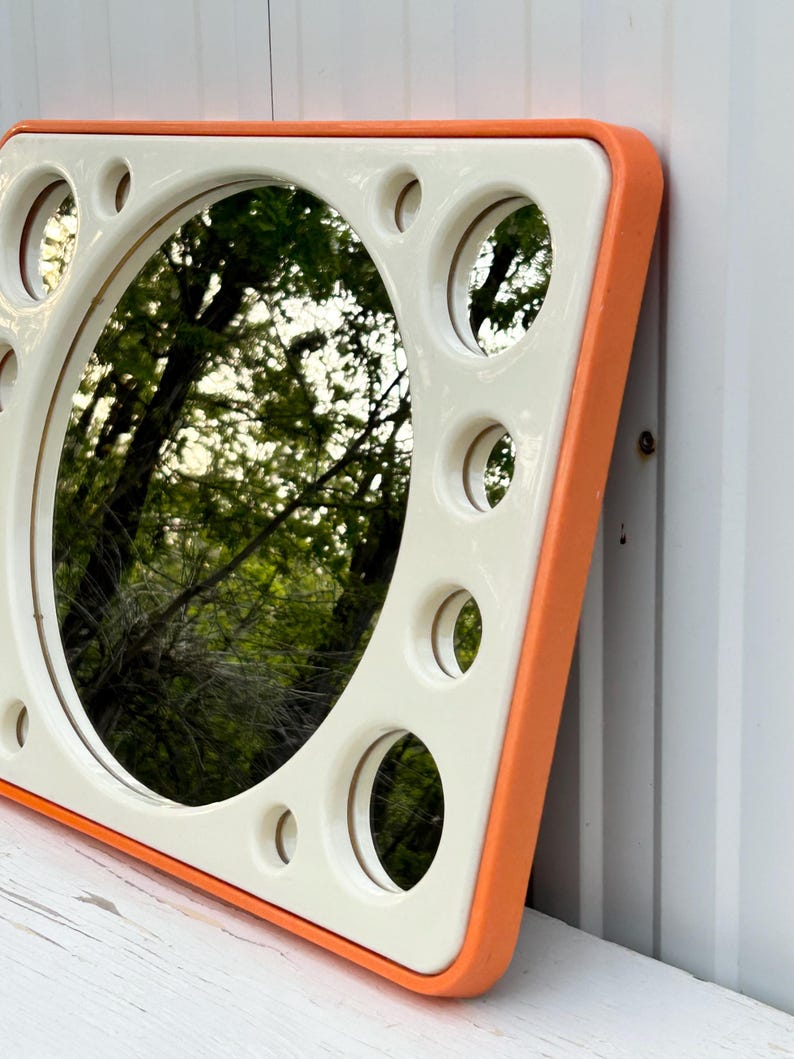 May include: A square mirror with a cream-colored frame and an orange border. The frame has several circular cutouts around the central mirror. The mirror reflects a view of trees and foliage.