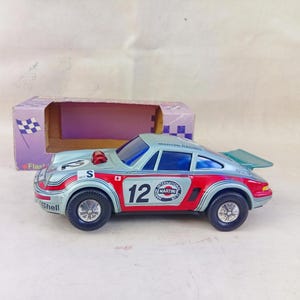 Può includere: Un'auto giocattolo argentata e rossa con il numero 12 sul lato. L'auto è una replica di una Porsche 911 e ha il logo Martini Racing sul lato. L'auto è in una scatola con una bandiera a scacchi sul lato.