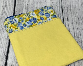May include: A yellow fabric case with a floral pattern at the top. The floral pattern features blue and yellow flowers on a yellow background. The case has a blue zipper and is likely for a tablet or e-reader.