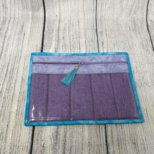 May include: A purple zippered pouch with a turquoise tassel. The pouch has a clear plastic front and a blue and white patterned fabric border.