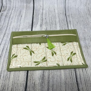 May include: A green zippered pouch with a white fabric lining featuring a dragonfly pattern. The pouch has a green tassel zipper pull.