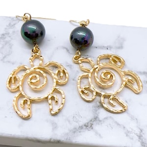 May include: A pair of gold-colored earrings featuring a sea turtle design. Each earring has a dark, iridescent pearl at the top and a turtle pendant with a spiral design. The earrings are displayed on a white and gray marble surface.
