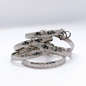 May include: A set of four silver bangles with a floral design. The bangles are stacked on top of each other and have a heart charm attached to one of them.