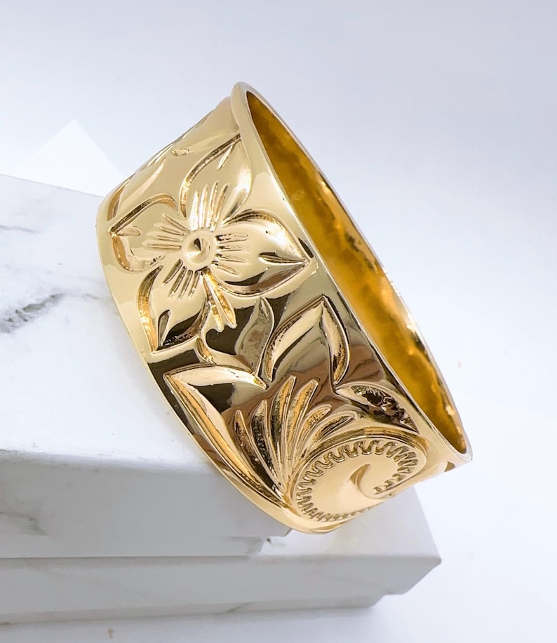 K&ouml;nnte beinhalten: Ein breiter, goldfarbener Ring mit einem gravierten Blumenmuster. Der Ring ist aus Metall und hat eine gl&auml;nzende Oberfl&auml;che.