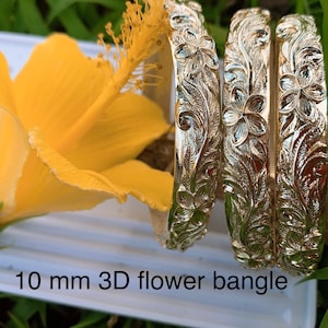May include: Three gold-tone metal bangles with a 3D flower design. The bangles are stacked on top of each other. The text "10 mm 3D flower bangle" is visible at the bottom of the image.