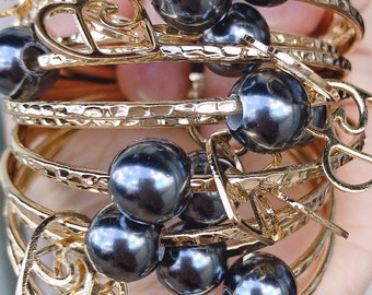 May include: A set of gold-toned bangle bracelets with black pearl accents. The bracelets feature a delicate, intricate design with heart-shaped cutouts.