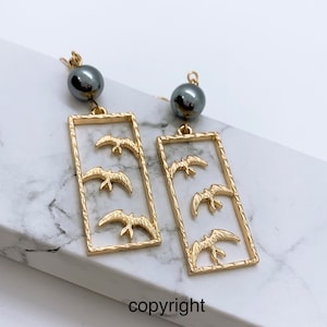 May include: Gold earrings with a rectangular frame and three flying birds inside. Each earring has a black pearl hanging from the top.