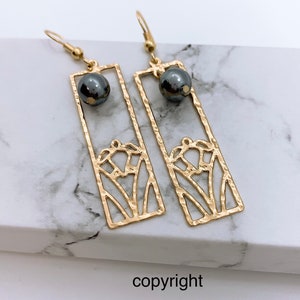 May include: Gold earrings with a delicate floral design and a black bead accent. The earrings are rectangular in shape and have a hammered texture.