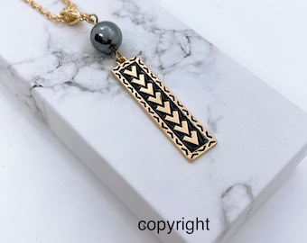 May include: Gold chain necklace with a black bead and a gold pendant with a black chevron pattern. The pendant is rectangular and has a gold border.