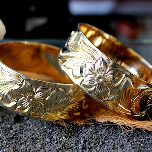 K&ouml;nnte beinhalten: Zwei goldfarbene Metallarmb&auml;nder mit aufwendigen Blumendesigns. Die Armb&auml;nder liegen &uuml;bereinander auf einer dunklen Oberfl&auml;che.