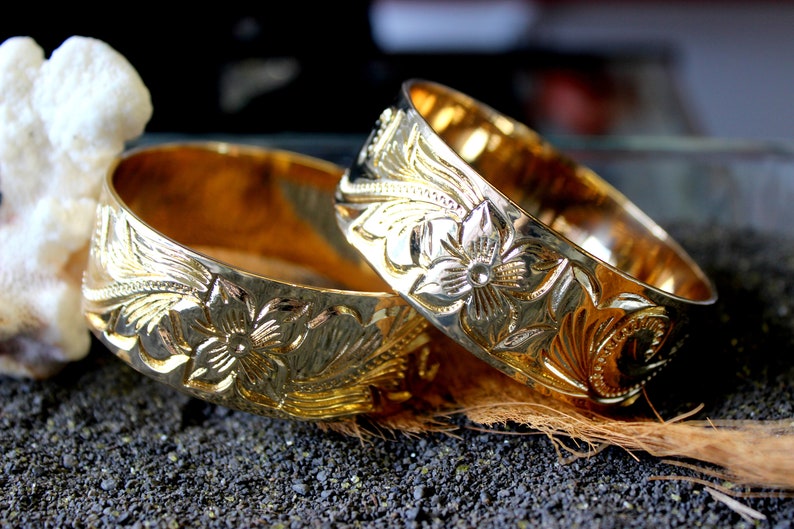 K&ouml;nnte beinhalten: Zwei goldfarbene Metallarmb&auml;nder mit aufwendigen Blumendesigns. Die Armb&auml;nder liegen &uuml;bereinander auf einer dunklen Oberfl&auml;che.