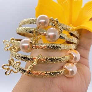 May include: Four gold-tone bangle bracelets with delicate floral designs and pearl accents. The bracelets are stacked on a hand.
