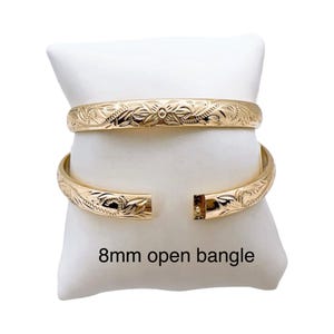 May include: Two gold-colored bangles with intricate floral patterns, one open and one closed, rest on a white pillow. The open bangle is labeled "8mm open bangle". The jewelry is displayed against a white background.