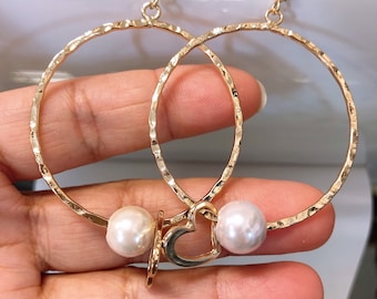 May include: Gold hoop earrings with a hammered texture and a heart charm. The earrings are accented with two white pearls.