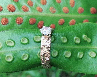 May include: A gold ring with a single diamond set in the center. The ring is on a green leaf with brown spots.