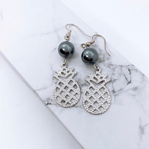 May include: A pair of silver pineapple earrings with black pearl accents. The earrings are dangling and have a delicate, intricate design.