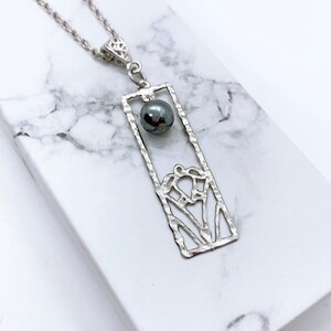 May include: A silver necklace with a rectangular pendant featuring a cut-out design and a dark grey bead.