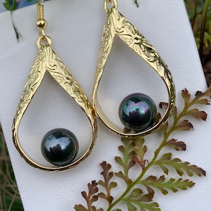May include: Gold-tone teardrop earrings with a floral design and a dark, iridescent pearl. The earrings have a hook closure. The jewelry is displayed on a white surface with green and brown foliage.