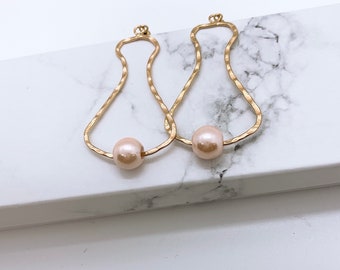 May include: Two gold-colored, hammered-texture earrings. Each earring features a teardrop-shaped frame with a light pink pearl bead at the base. The earrings are displayed on a white and gray marble surface.