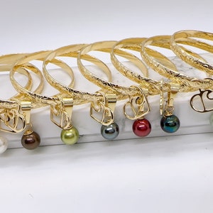 May include: Eight gold bracelets with engraved designs and heart-shaped charms. Each bracelet has a different colored pearl attached to the charm.